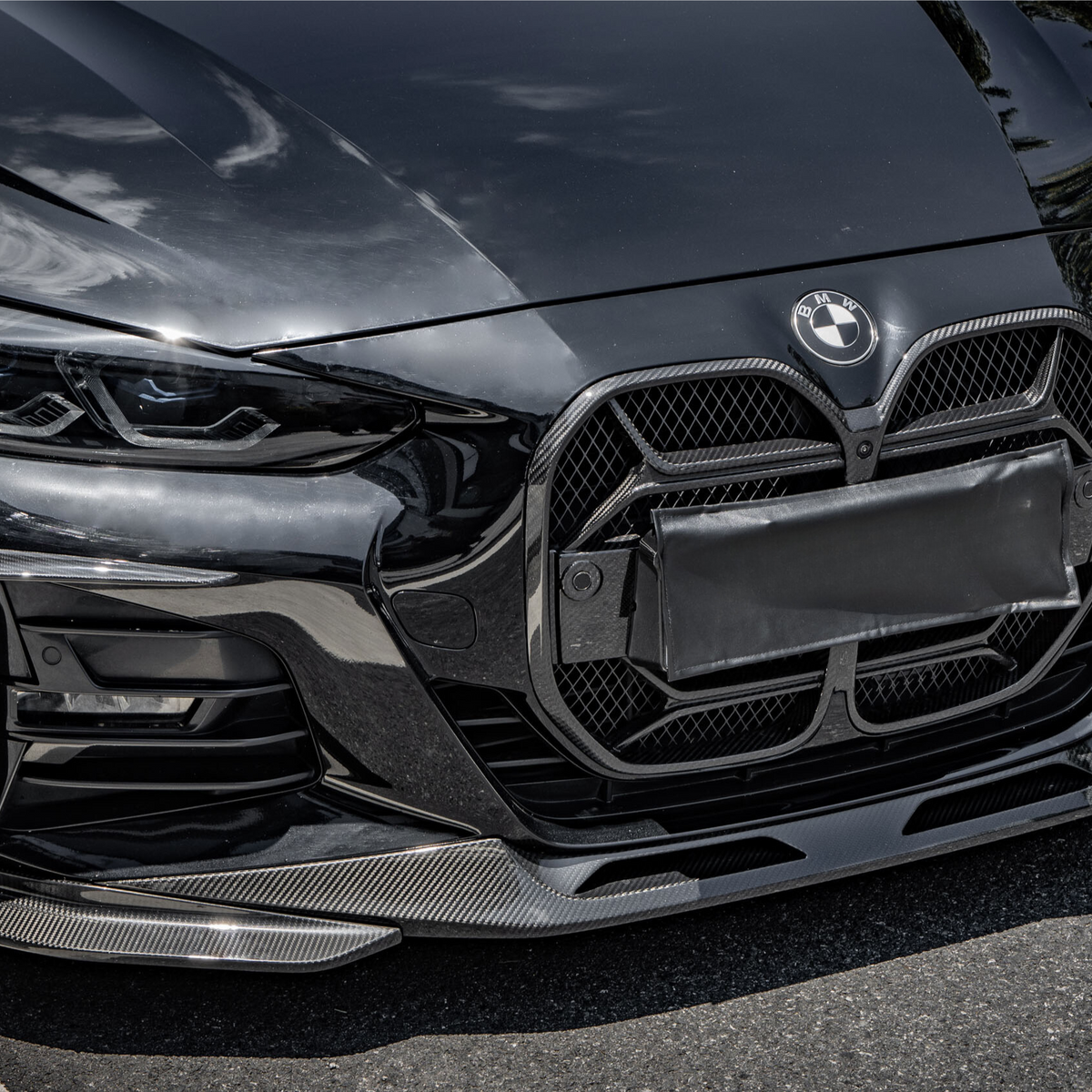 A black luxury car with a prominent grille and sleek headlights is parked on a road. Sunlight reflects off its shiny surface, highlighting its modern design. The BMW logo is visible.