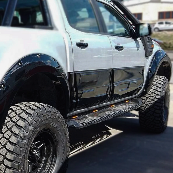 A lifted, black-and-white pickup truck is parked on a road, featuring large off-road tires and side steps with orange reflectors. It’s set against a blurred background.