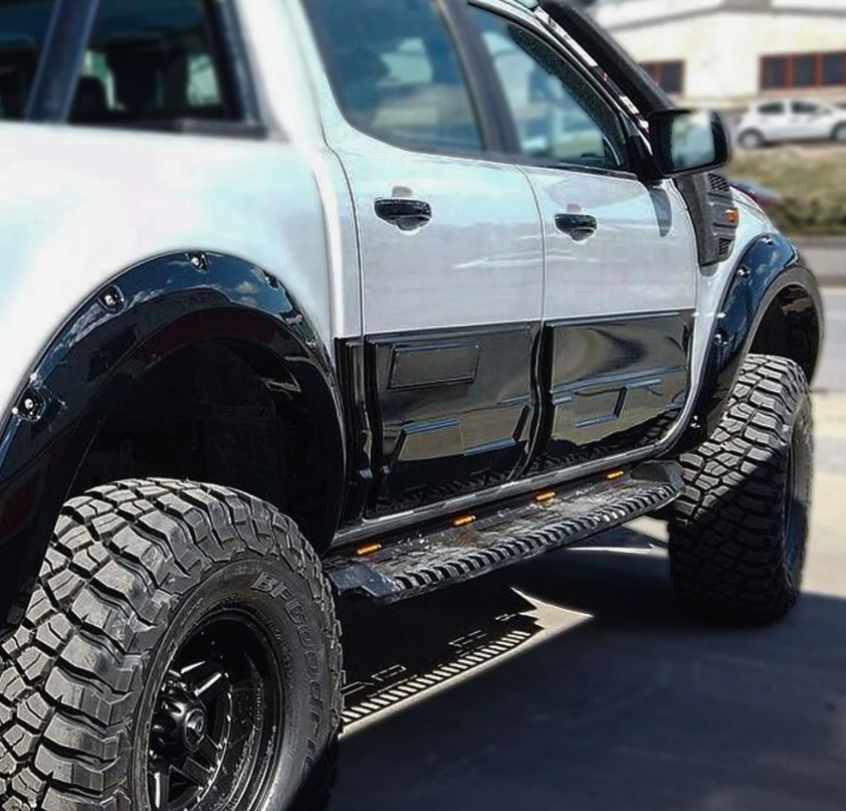 A lifted, black-and-white pickup truck is parked on a road, featuring large off-road tires and side steps with orange reflectors. It’s set against a blurred background.