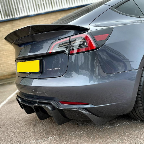 A sleek gray car with a "DUAL MOTOR" badge and a prominent rear spoiler is parked on an asphalt surface near a brick wall. The license plate is yellow.