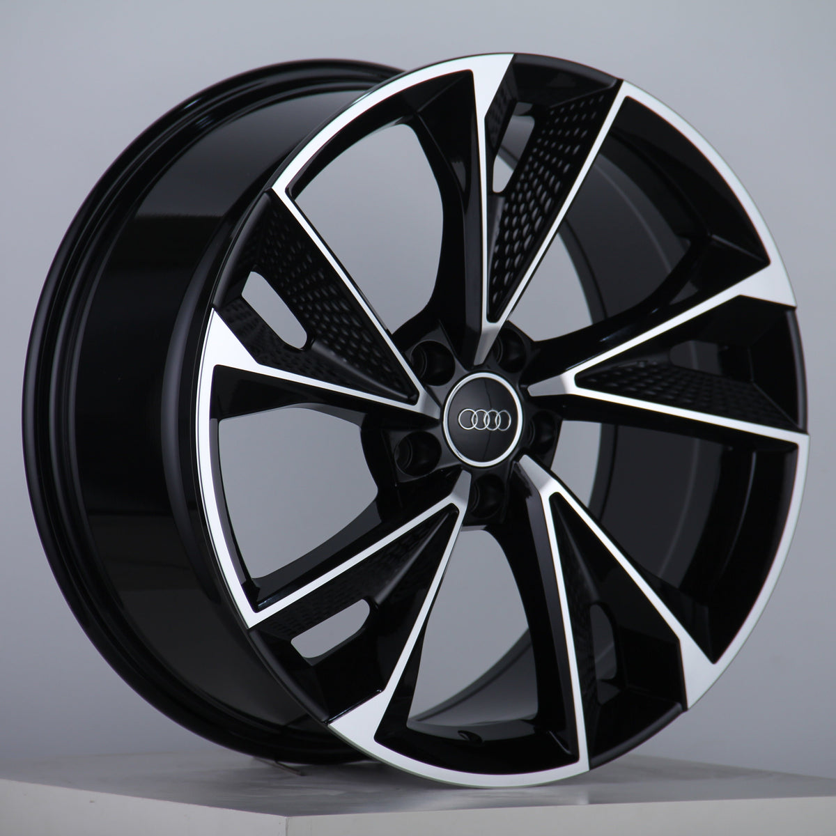 A black and silver alloy wheel displays its intricate, angular design with five spokes. The Audi logo is centered. It is set against a plain gray background, resting on a white surface.