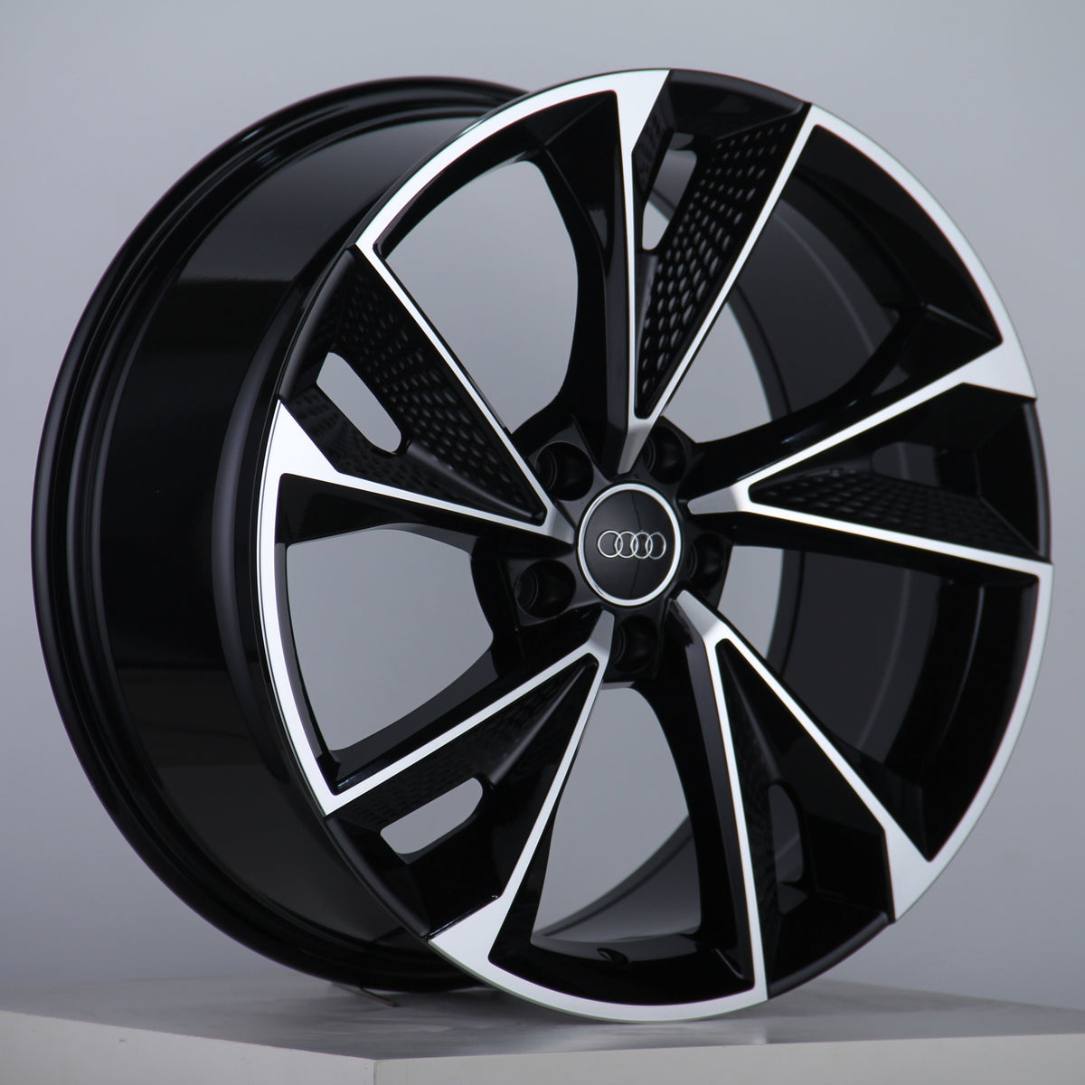 A black and silver alloy wheel with a five-spoke design features the Audi logo at the center, displayed on a simple white platform against a plain gray background.