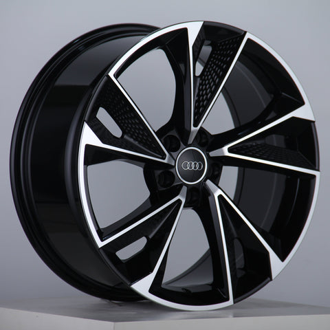 A black and silver alloy wheel with a five-spoke design features the Audi logo at the center, displayed on a simple white platform against a plain gray background.