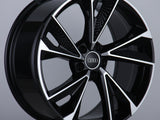 A black and silver alloy wheel featuring the Audi logo at its center, positioned upright on a gray surface in a neutral setting.