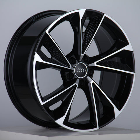 A sleek, black and silver alloy wheel with the Audi logo stands on a grey surface against a neutral background, showcasing a modern, geometric design.