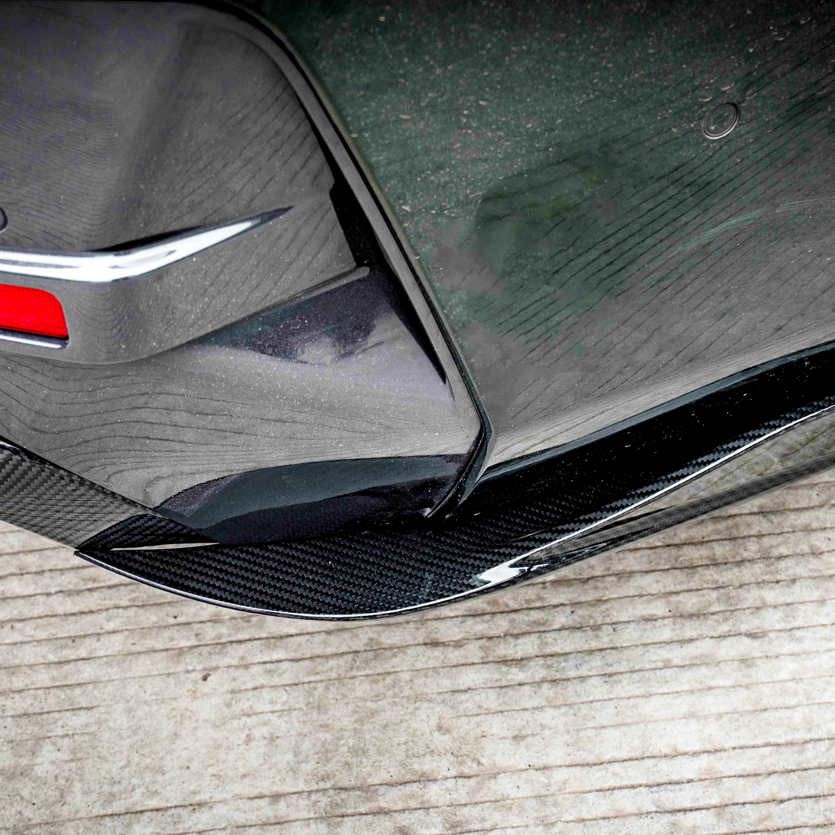 Car bumper features a red reflector and carbon fiber-like trim, positioned near a rear tire. The vehicle is parked on a grooved concrete surface.