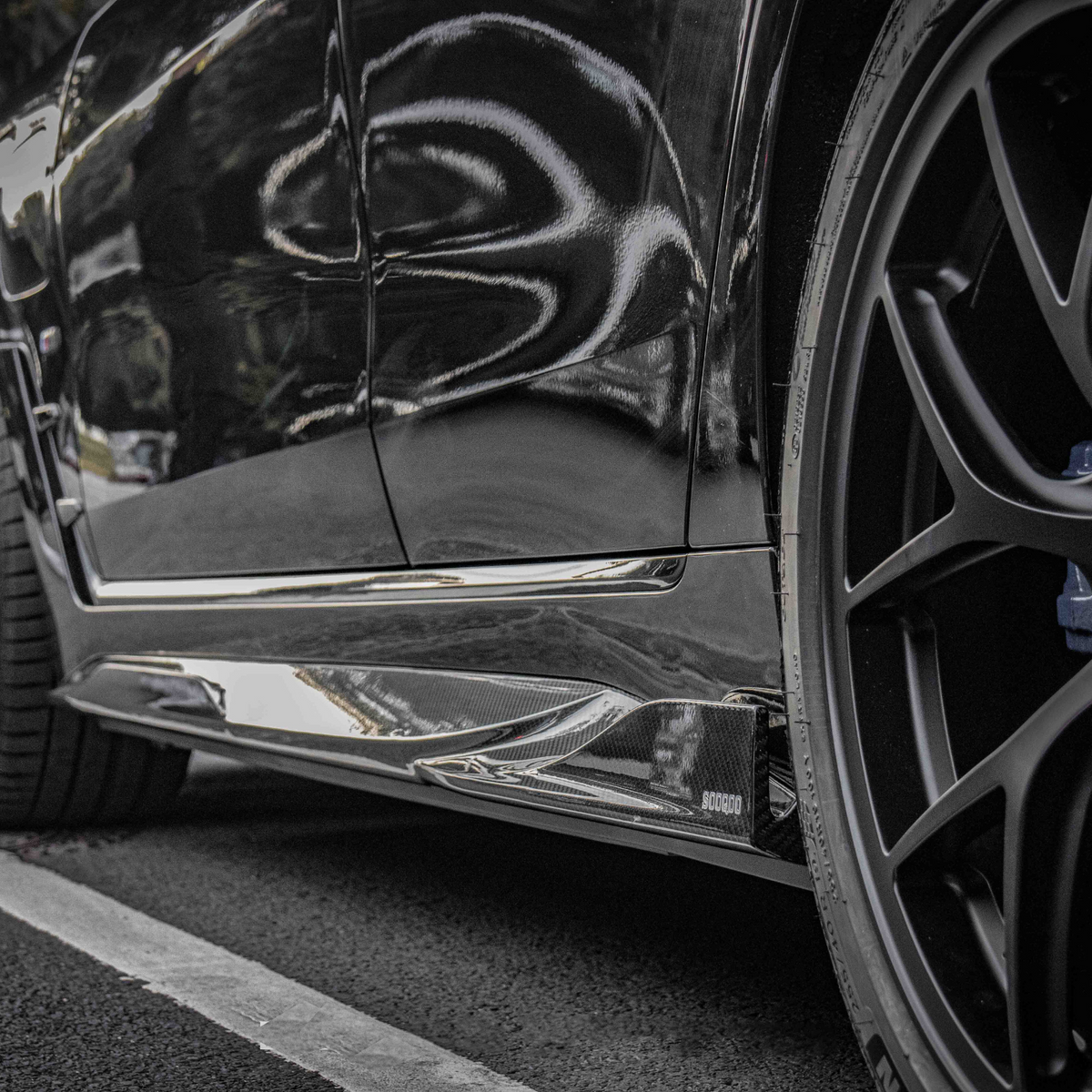 A black car's side view, showing glossy bodywork and a detailed wheel with a BMW logo, parked on asphalt near trees. Visible text includes "SOCODA."