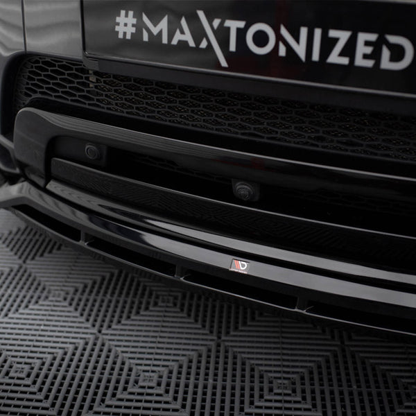 A black car's front bumper displays the text "#MAXTONIZED" on a license plate with a hexagonal grille, parked on a patterned rubber mat.