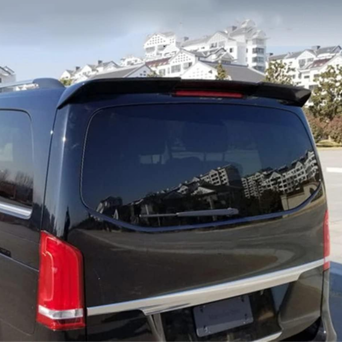 A black vehicle with tinted windows is parked, reflecting nearby white buildings with sloped roofs. The scene is set in a sunny, open outdoor area.