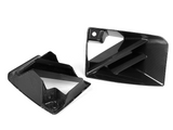 Two black, angular carbon fiber car parts are positioned on a white background. One lies flat, showing its patterned surface, while the other stands upright, revealing defined geometric angles.