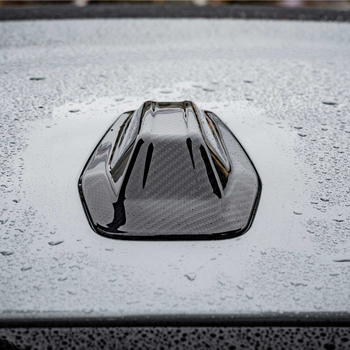 A black, shark fin car antenna rests on a gray, wet car roof, with raindrops scattered across the surface creating a textured appearance.