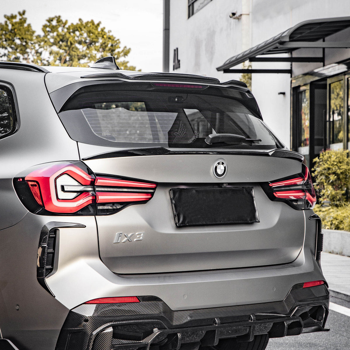 A BMW iX3 parked on a street, featuring prominent taillights and a sleek rear design. It's positioned near a modern white building with green foliage in the background.