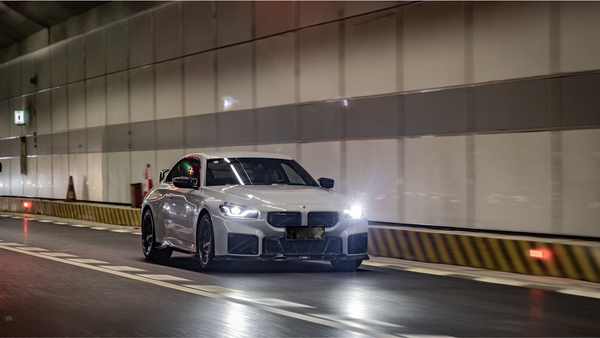 A white sports car speeds through a well-lit tunnel, headlights illuminating the path. The tunnel's striped walls and traffic cones provide a sense of motion and urban environment.