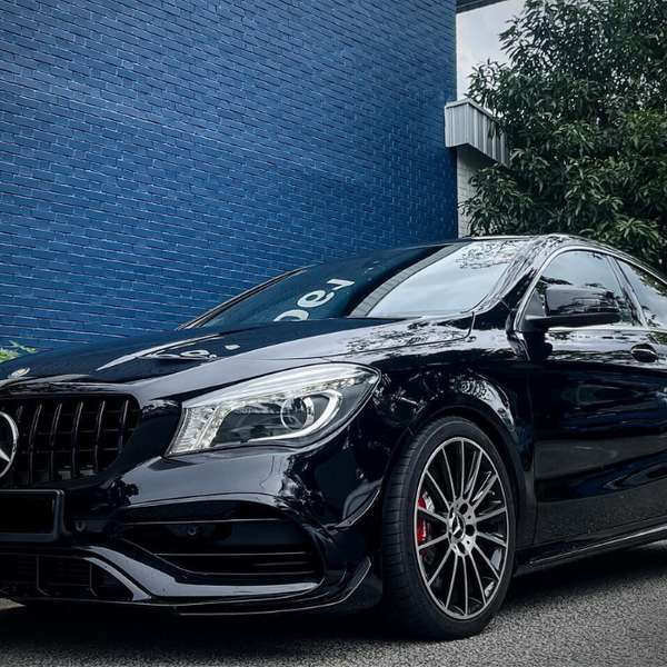 A black Mercedes-Benz car is parked on a street beside a blue brick wall, with greenery nearby and a tree overhead, conveying a sleek, urban environment.