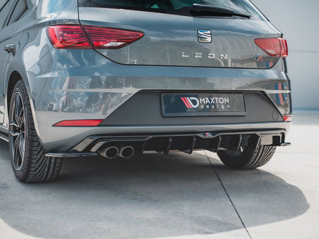 A gray hatchback car, parked on a smooth surface, displays a sporty rear bumper with dual exhausts. The license plate reads "MAXTON design," surrounded by a bright outdoor environment.