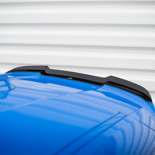 A blue car roof features a black spoiler. Set against a corrugated white background, the reflective surface of the car adds dynamic lines to the composition.