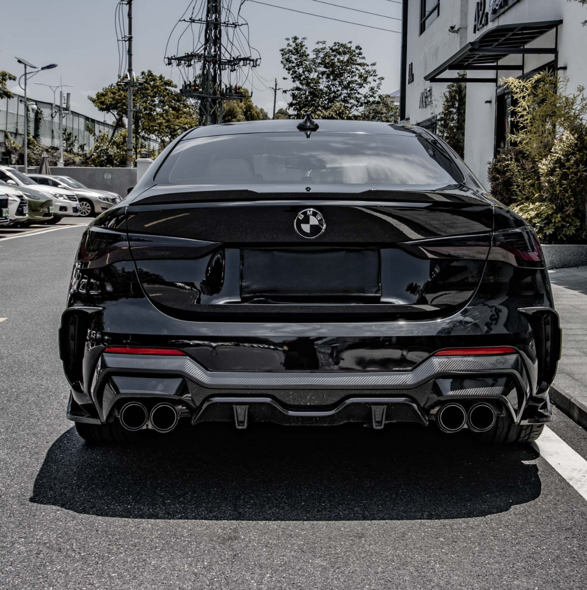 Shiny black sedan parked, features quad exhausts and sleek tail design; in an urban parking area with surrounding cars, electrical poles, greenery, and buildings in the background.