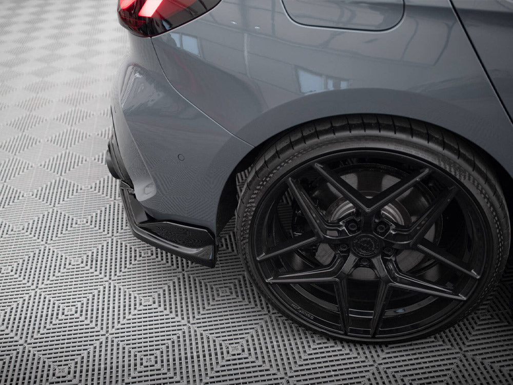 A car's rear wheel and bumper, featuring a sleek black alloy and low-profile tire, rests on a patterned grey floor with diamond shapes in an indoor setting.