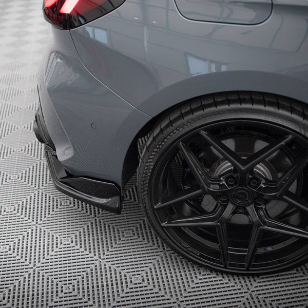 A car's rear wheel and bumper, featuring a sleek black alloy and low-profile tire, rests on a patterned grey floor with diamond shapes in an indoor setting.