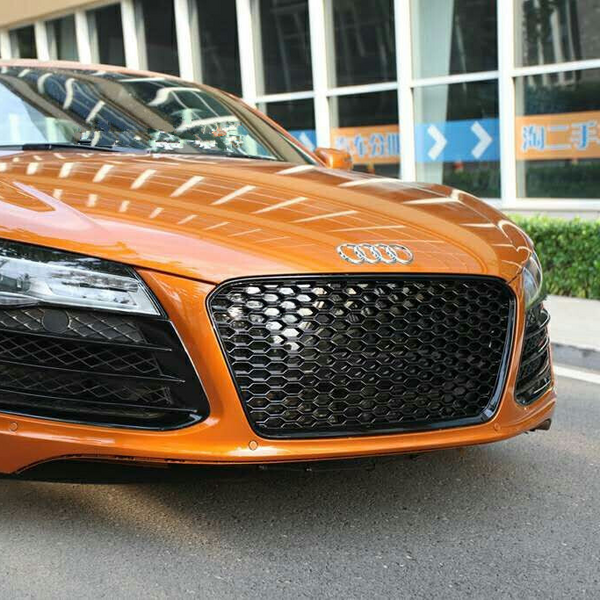 Orange sports car parked on a street, featuring a distinctive honeycomb grille and prominent Audi emblem. Background includes windows and a hedge, with Chinese text on visible signs.