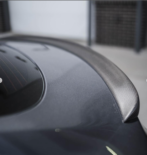 A sleek carbon fiber spoiler rests on a gray car's trunk, reflecting light subtly. The car is parked indoors against a neutral backdrop with lattice panels in the distance.