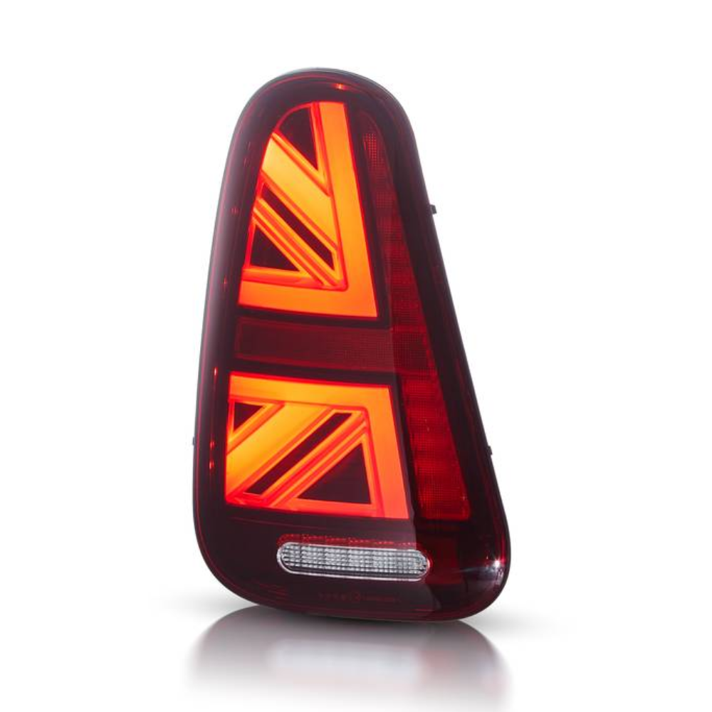 A red automotive tail light with illuminated chevron patterns is positioned upright against a plain white background. It features a horizontal white reflector at the bottom.