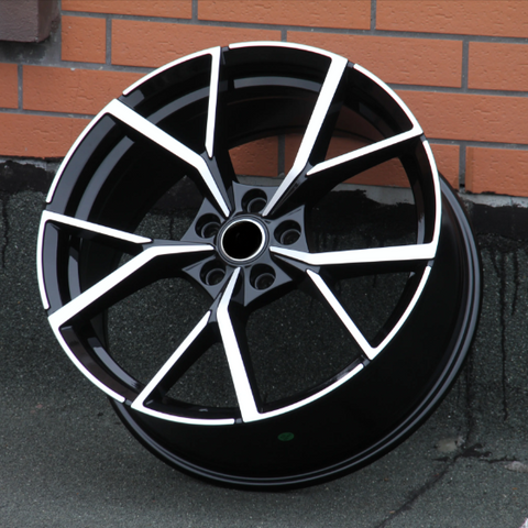 A black alloy wheel with a modern, angular design featuring silver accents rests against a brick wall on a textured concrete surface.