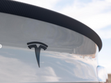 The rear of a white car displays the Tesla logo, with a carbon fiber spoiler against a partly cloudy sky.
