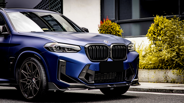 A blue SUV with a visible front grille and emblem is parked on a street. The scene includes surrounding greenery and a building with black-framed windows.