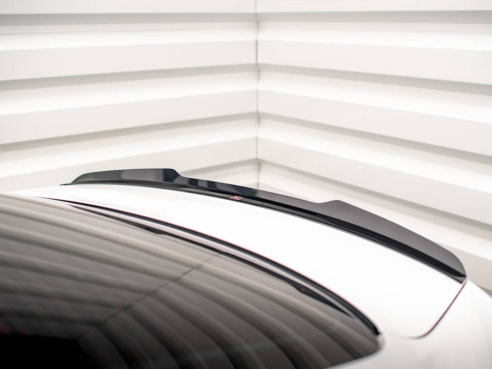 A black spoiler is positioned on a white car trunk, with a sleek design. The setting is a room with white, horizontally-lined walls, adding a modern aesthetic.
