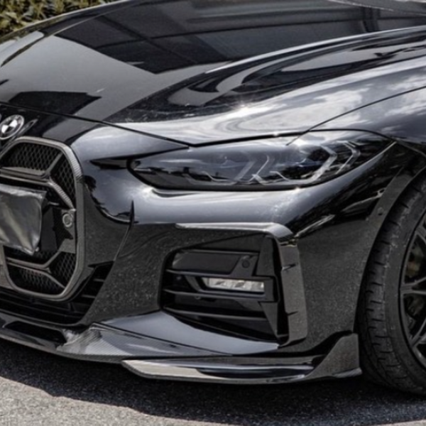 A sleek, black car displays its front grille and headlight assembly while parked on an outdoor surface. The wheel features a detailed spoke design, with greenery visible in the background.