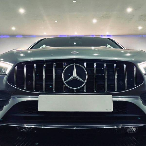 A silver car with a prominent grille and illuminated headlights is parked indoors under bright ceiling lights, creating a sleek and modern atmosphere. A Mercedes-Benz logo is centered on the grille.