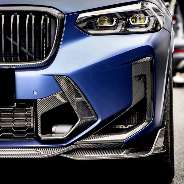 A luxury car's front, featuring a sleek, blue grille with carbon fiber accents, is parked beside a road. In the background, a silver car's tailgate is open with items inside.