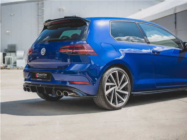 A blue hatchback car is parked on a concrete surface outside a building. The license plate reads "DVUXITION DES UAE." The car features dual exhausts and sporty wheels.