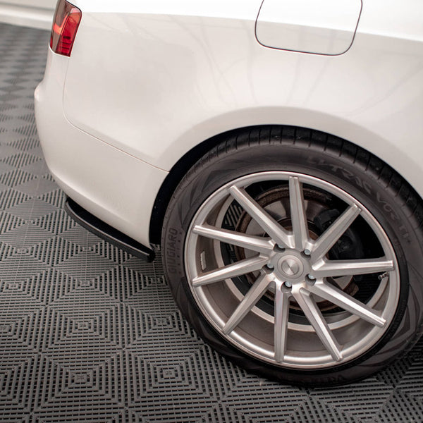 A white car's rear wheel with a silver alloy rim is stationary on a textured gray garage floor, showing partial tail light and fuel door.
