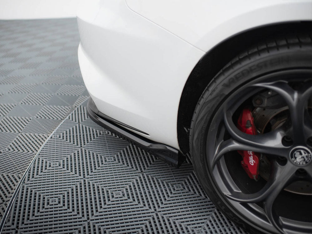 A white car's rear bumper is parked on a patterned grid floor. The black alloy wheel features red brake calipers labeled "Alfa Romeo." The tire brand reads "VREDESTEIN."
