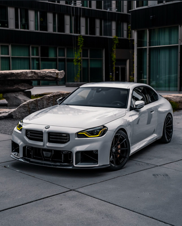 A sleek white sports car with yellow headlights is parked on a concrete surface, surrounded by modern glass buildings and large stone sculptures. BMW logo visible on the grille.