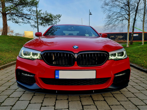 A red BMW car is parked on a cobblestone driveway, framed by grassy areas and trees with overcast skies in the background. The license plate reads "IRL."