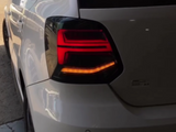 A white car's rear section with illuminated tail lights is parked near a building, casting reflections on its glossy surface.
