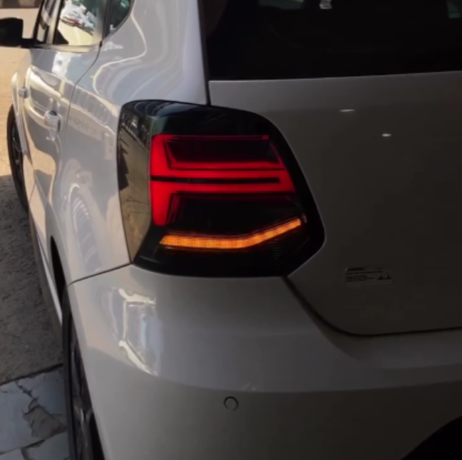 A white car's rear section with illuminated tail lights is parked near a building, casting reflections on its glossy surface.