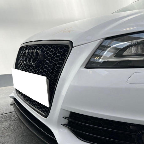 A white car's front features a prominent black grille with a logo and sleek headlight, situated on a paved surface against a blurred, grey background.