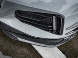 A car's front bumper, featuring sleek black grills and carbon fiber accents, is covered in rain droplets, situated on a wet asphalt surface with white road markings.
