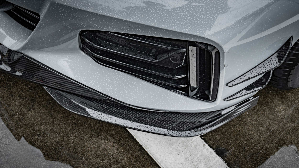 A car's front bumper, featuring sleek black grills and carbon fiber accents, is covered in rain droplets, situated on a wet asphalt surface with white road markings.