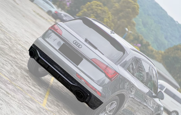 A gray Audi SUV is parked on a concrete lot, surrounded by other cars. Trees and hills create a natural backdrop. The rear badge features the Audi logo.