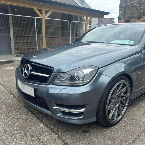 A silver Mercedes car is parked on a concrete surface near a metal fence and wooden structure. The car's windshield displays documents. The setting is an outdoor area adjacent to buildings.