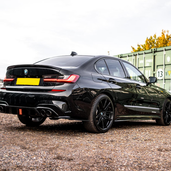 A sleek black car is parked on a gravel lot near green shipping containers. The car's rear features twin exhausts and shiny black rims, and the background has trees and buildings.