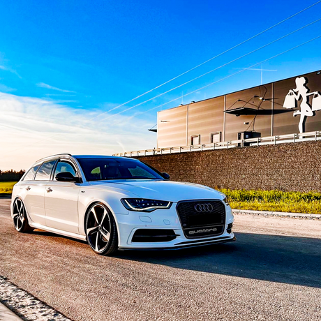 A white Audi station wagon parked on a sunny road near a modern building with a mural of a woman holding shopping bags. The license plate reads "QUATTRO".