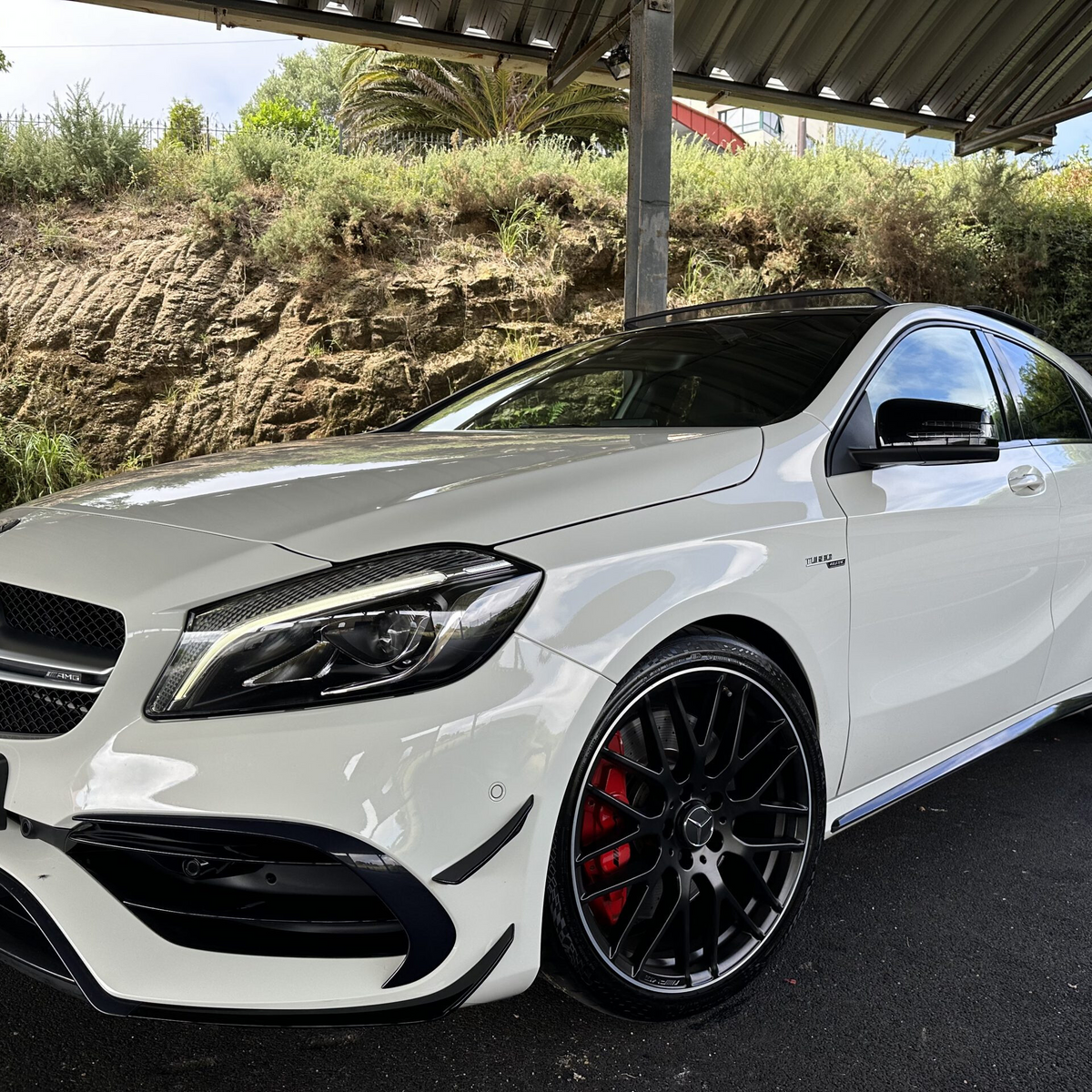 A white Mercedes-Benz car, parked under a metal shelter, features black alloy wheels and is set against a background of greenery and a rocky wall.