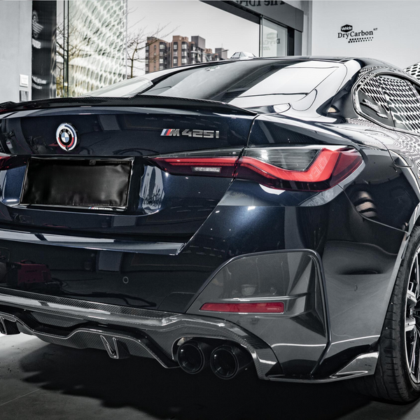 A black BMW M425i car is parked indoors, reflecting light on its glossy surface. Nearby, a tool cabinet and bottle indicate a workshop environment. "DryCarbon" branding is visible on the wall.