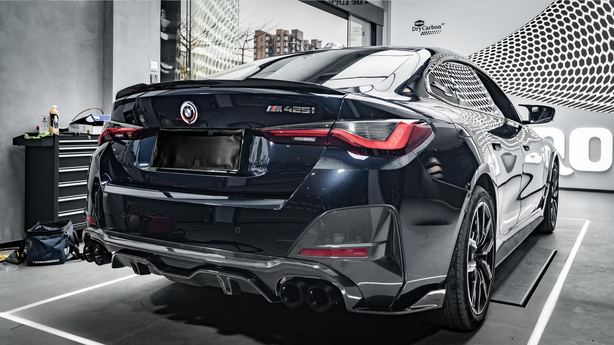 A black BMW M425i car is parked indoors, reflecting light on its glossy surface. Nearby, a tool cabinet and bottle indicate a workshop environment. "DryCarbon" branding is visible on the wall.